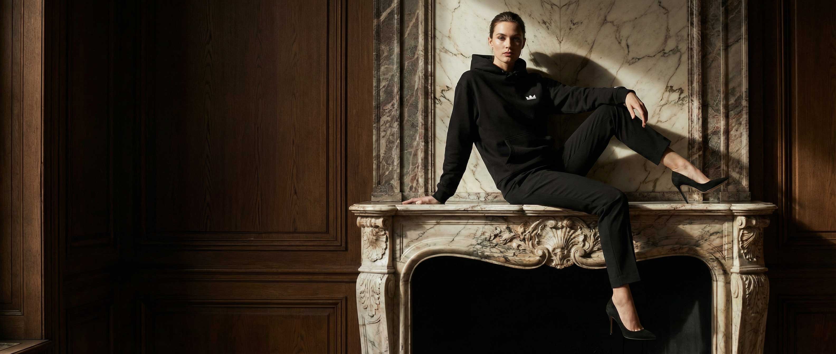 Model in a black GG UNLTD hoodie posed on an ornate marble fireplace in a dark wood-paneled room